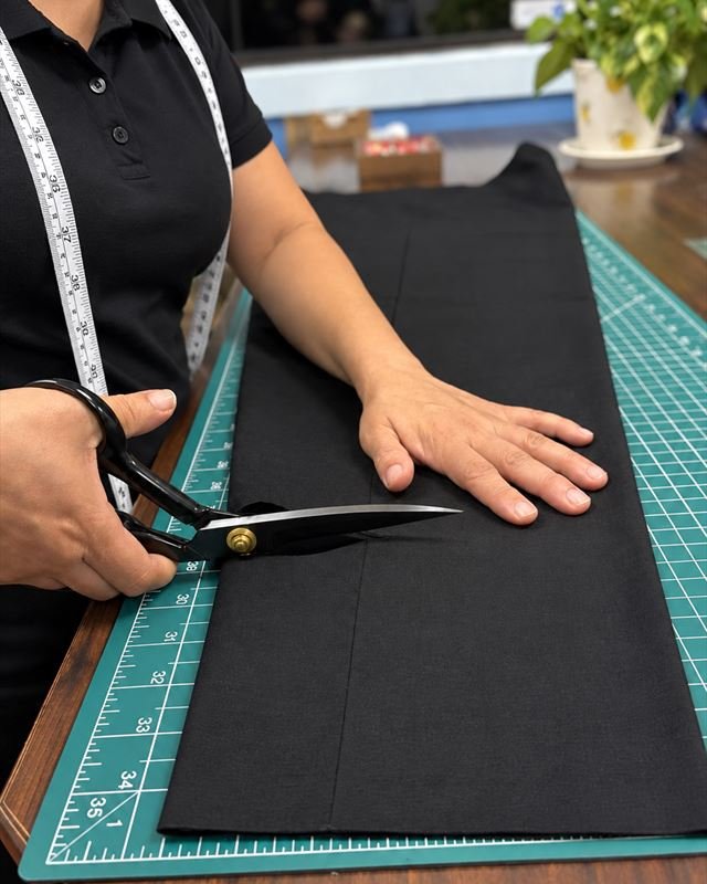 A tailor with a tape measure around her neck cutting black fabric on a cutting mat.