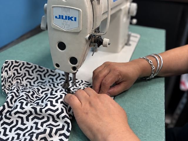 Hands guiding a patterned fabric under the foot of a Juki sewing machine at the shop.