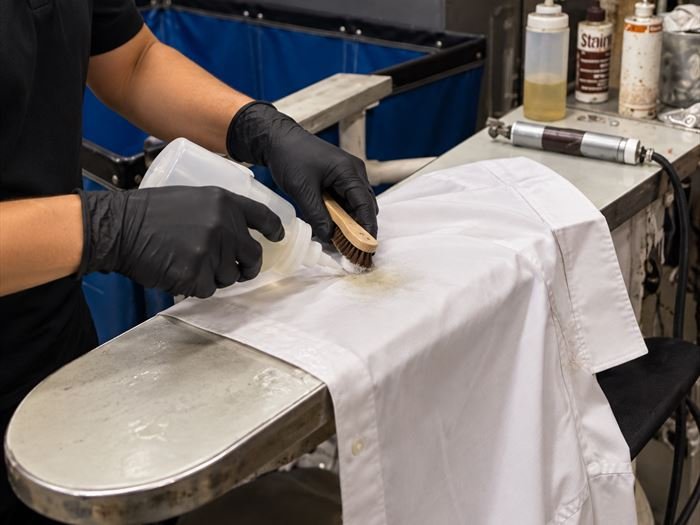 A gloved hand working spotting solution into a white shirt collar with a soft brush.